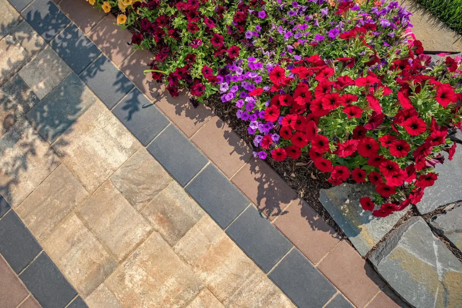 A newly paved black asphalt driveway curves past a brick house with green lawns and landscaping, bordered by stone edging and surrounded by tall trees.