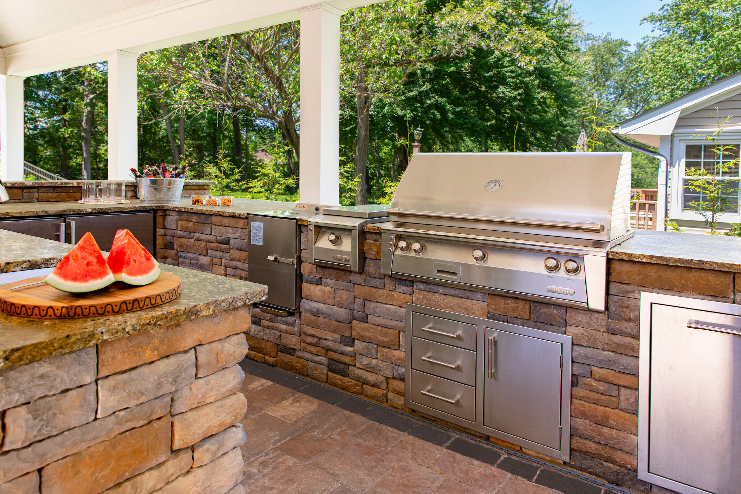A newly paved black asphalt driveway curves past a brick house with green lawns and landscaping, bordered by stone edging and surrounded by tall trees.