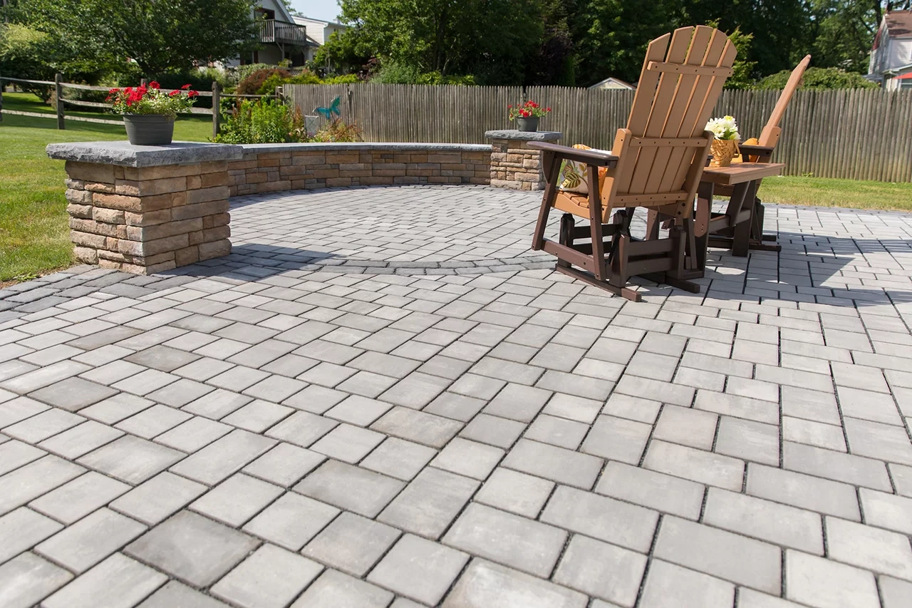 A newly paved black asphalt driveway curves past a brick house with green lawns and landscaping, bordered by stone edging and surrounded by tall trees.