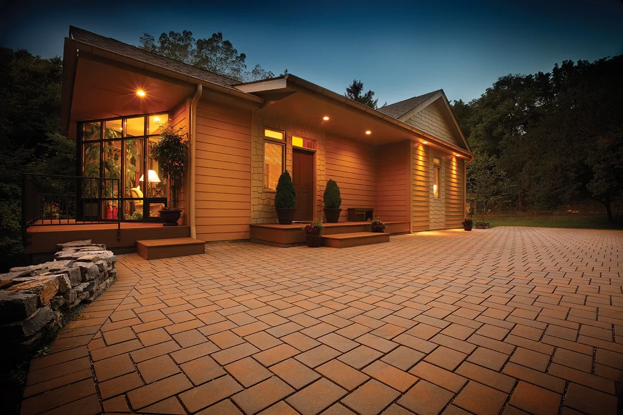 A newly paved black asphalt driveway curves past a brick house with green lawns and landscaping, bordered by stone edging and surrounded by tall trees.
