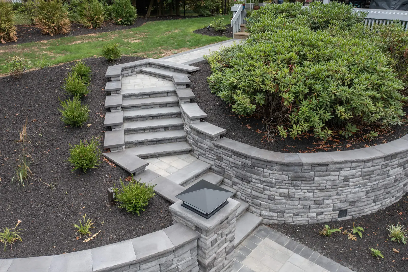 A newly paved black asphalt driveway curves past a brick house with green lawns and landscaping, bordered by stone edging and surrounded by tall trees.